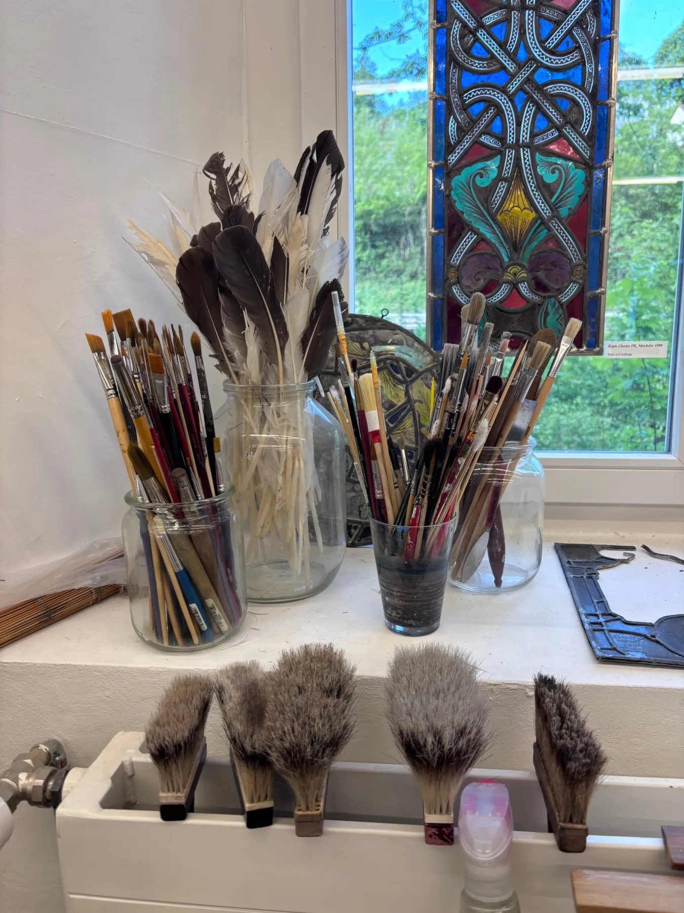 Pinsel, Federn und Werkzeuge vor einem farbigen Glasfenster im Atelier