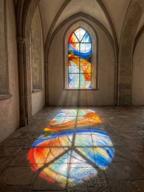 Ein farbiges Glasfenster in einem gotischen Raum wirft sein Licht in Orange, Blau und Rot auf den Steinboden